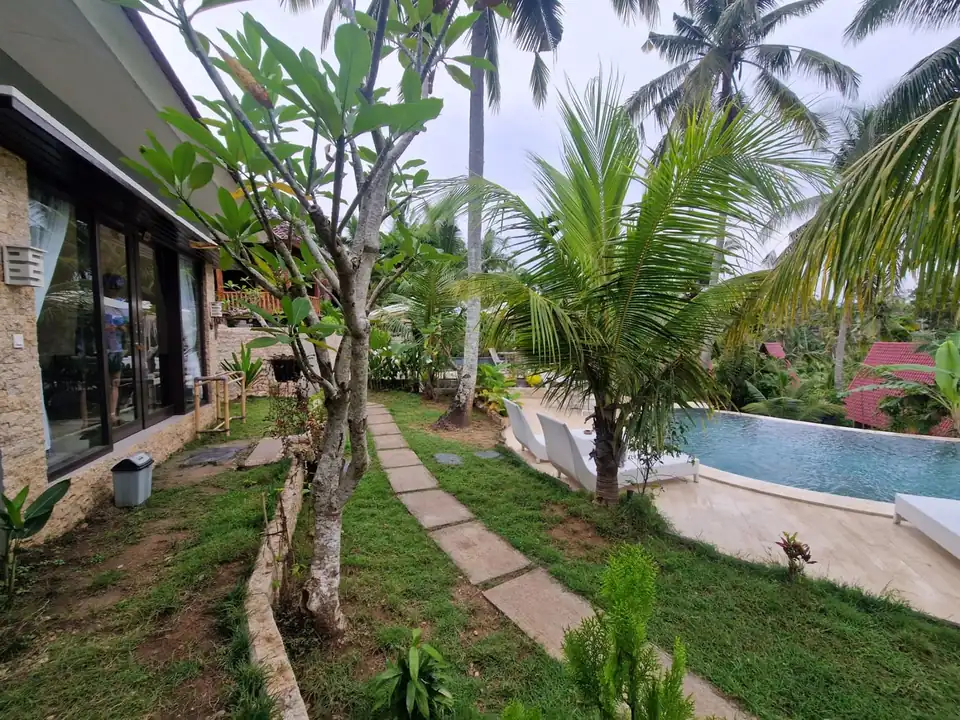 A stone path through a garden past villa rooms with glass doors and a pool beyond
